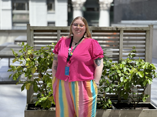 Woman wearing a bright pink top and rainbow pants, standing and smiling at the camera.