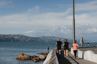People running on the side of the road with the harbour in the background.