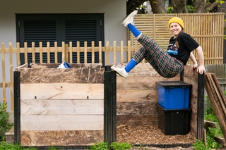 A woman wearing a yellow beanie and a black tshirt with checkered pants leaning on the side of a compost bin.