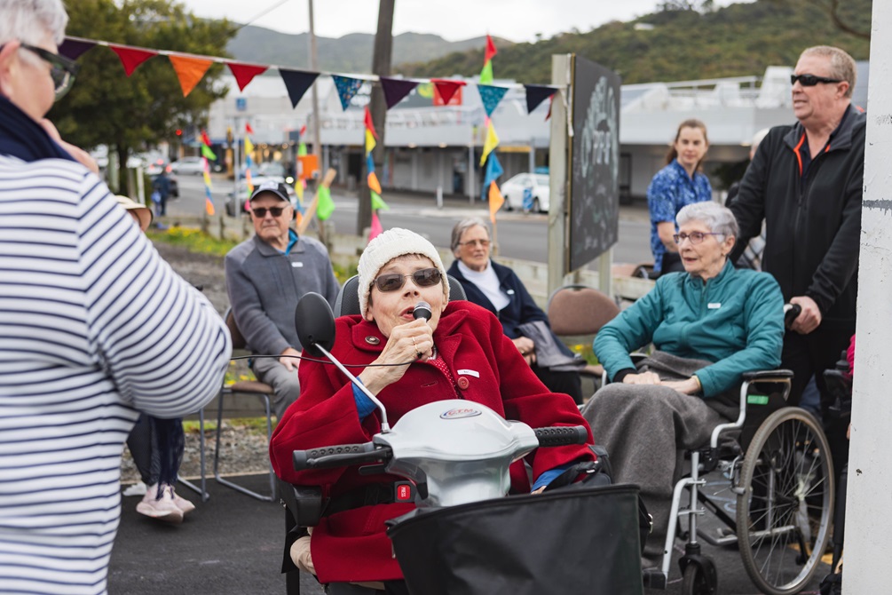 Huntleigh Retirement Village Valerie Smith speaks holding a microphone as she sits in her wheelchair wearing a woolly hat and read coat, in a suburban street setting with bunting decorations and other elderly folk.