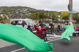 A very large green ribbon is cut by a woman wearing a read coat as she sits in a wheelchair on a pedestrian crossing. Onlookers stand on the roadside under street decorations.