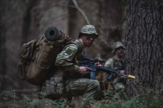 A man in his army gear during a military exercise, crouching down on the ground holding a weapon and wearing a large pack.