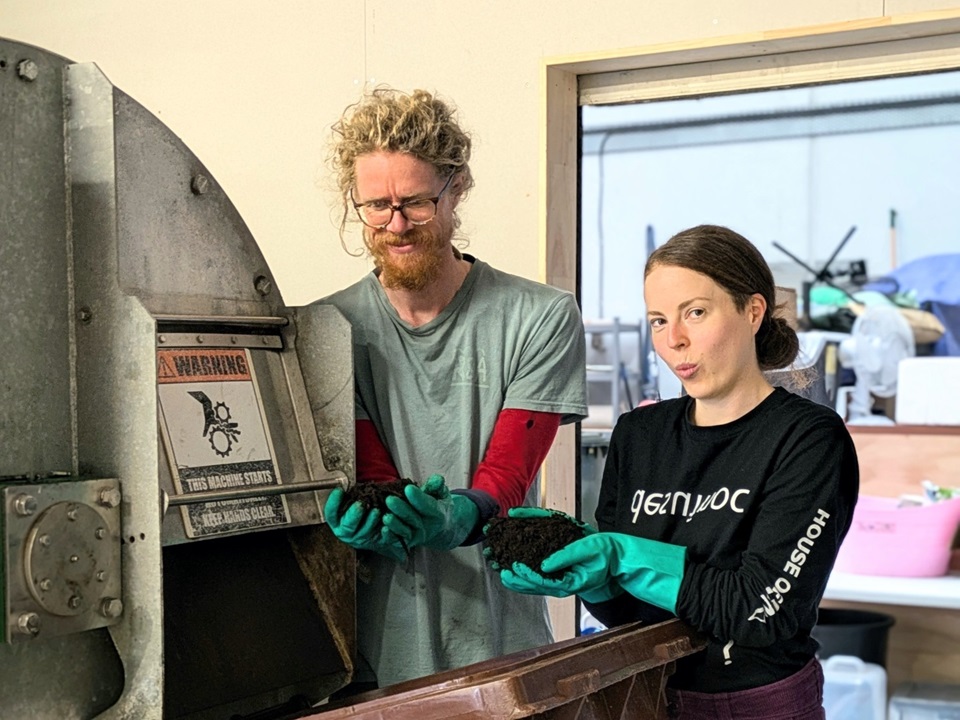 A man and a woman standing next to a composting machine, collecting compost in their hands.