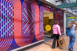 Two people walking down the street with artwork in the windows of the shops next to them.