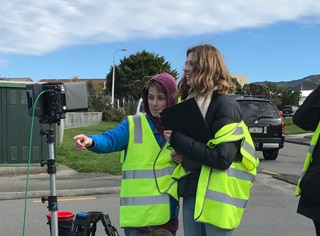 A young boy in a yellow high vis jacket, pointing ahead. He is standing next to an older woman who is holding a clipboard.