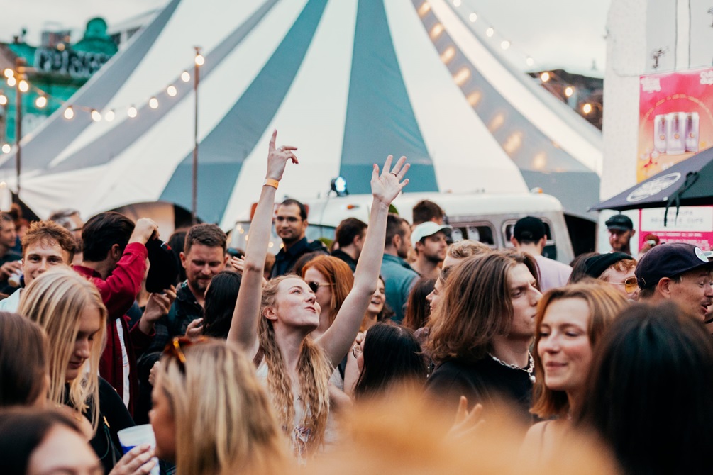 In the background there is a blue and white tent top, and infront there is a crowd of people dancing and socialising. 