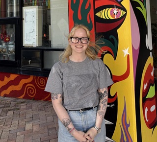 Person wearing a grey tshirt and blue jeans standing infront of a brightly painted pillar.