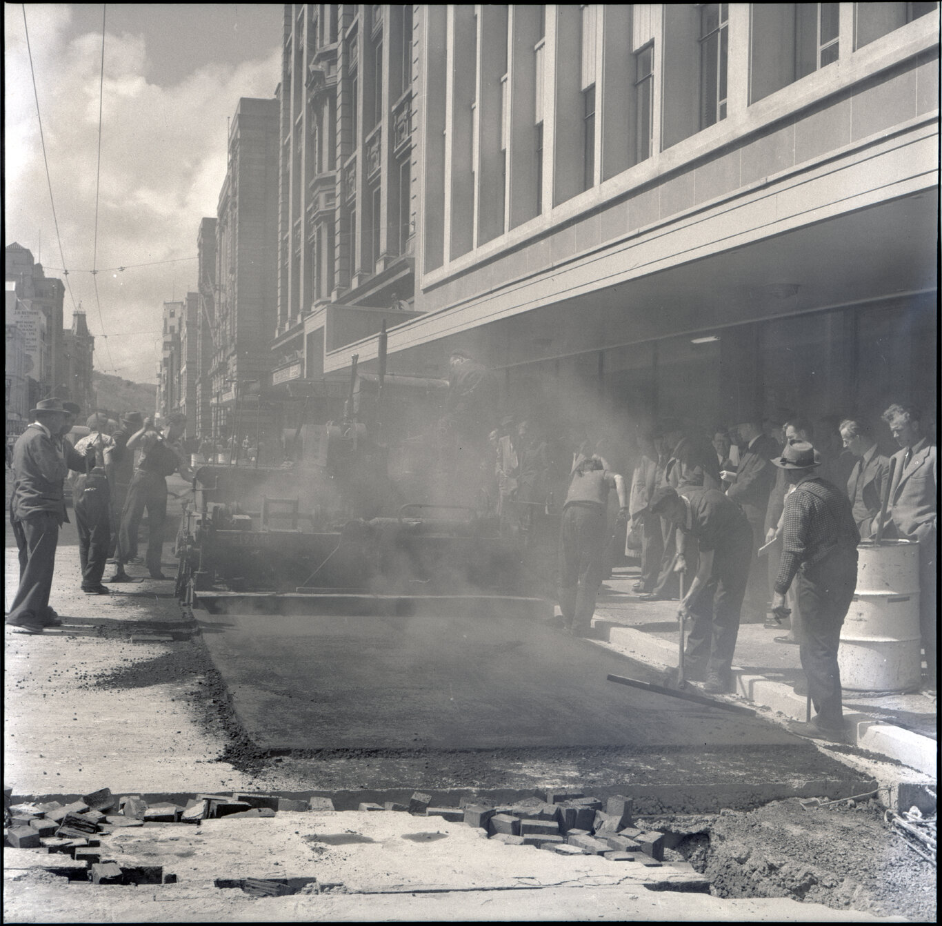 Various stages of asphaltic road surfacing by Council workmen, Lambton Quay end of Featherston Street. People passing by have stopped to watch work in progress. Trolly buses in background.