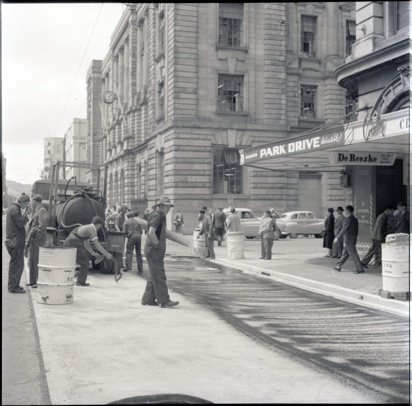 Various stages of asphaltic road surfacing by Council workmen, Lambton Quay end of Featherston Street. People passing by have stopped to watch work in progress. Trolly buses in background