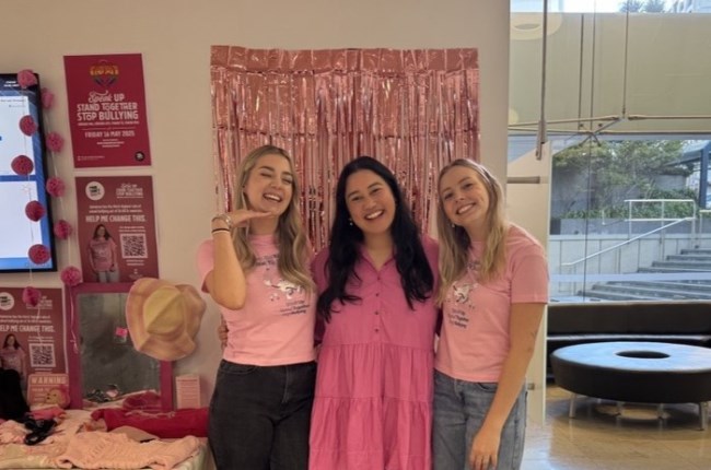 Three women standing together infront of a pink wall.