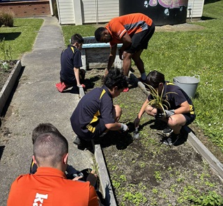 Young students planting in the garden with two adult supervisers wearing orange tshirts.