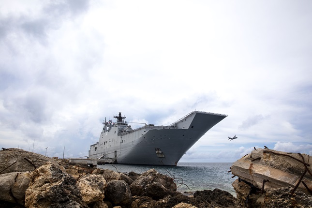 HMAS Canberra front at sea with rocks in foreground and plane in background.