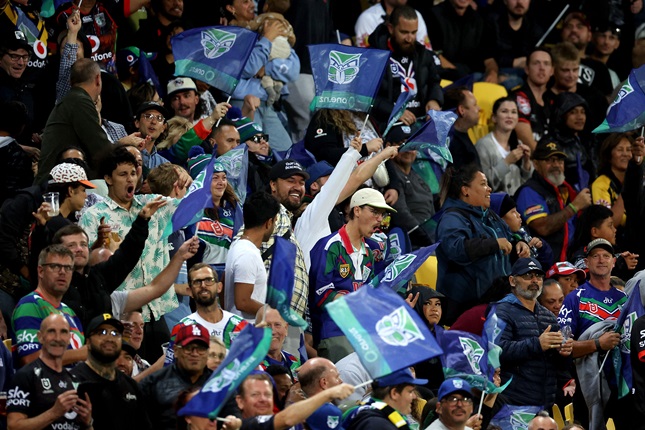 Fans in stands supporting Warriors game with flags and branded clothing.