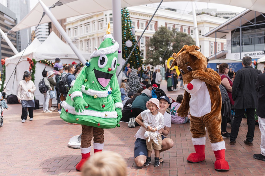 People dressed as a Christmas tree and a reindeer entertain children at an outdoor event.