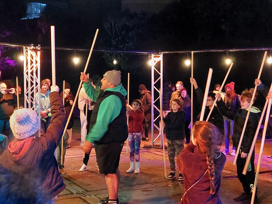 A mix of people holding large sticks and learning to play a traditional Māori game.