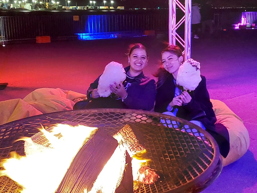 Two young women sit on bean bags in front a roaring brazier. They are both holding large cotton candy sticks.