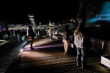 Observers stand and watch a memorial wall, where messages are being projected onto the side of a building.