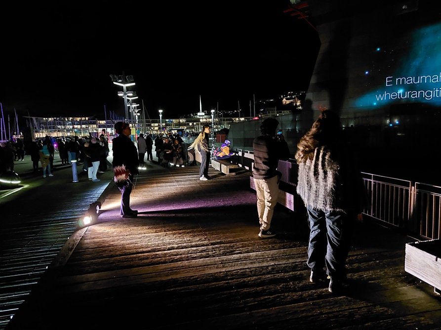 Observers stand and watch a memorial wall, where messages are being projected onto the side of a building.