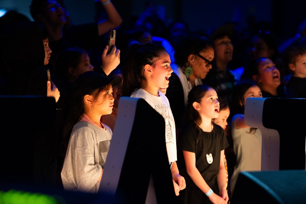 Three delighted children stand at the front of crowd, watching a performance.