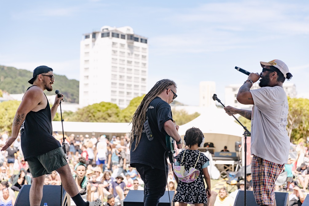 A band performs on stage with a large crowd on the grass in the background.