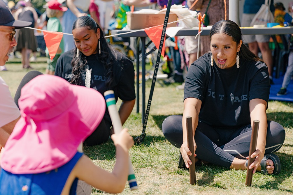 A group of people of various ages playing a game involving holding two wooden sticks.
