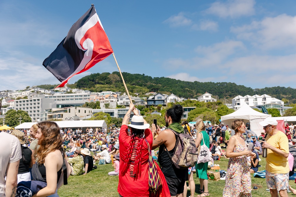 A couple in the event crowd wave a red, white and black Tino Rangatiratanga flag against a blue sky.