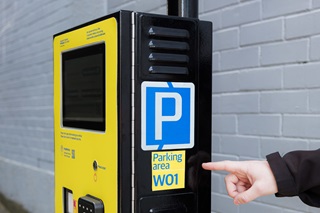 Pay by Plate parking meter with a person's hand pointing to parking area 'W01' on the side of the meter.