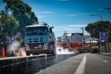 A layer of fresh bitumen on a road surface. A work crew and trucks are visible in the background.