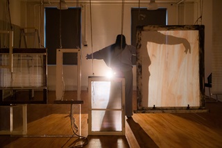 An exhibition showing a person in the centre with a light behind them and their shadow displaying on wooden frames and glass hanging from the roof.